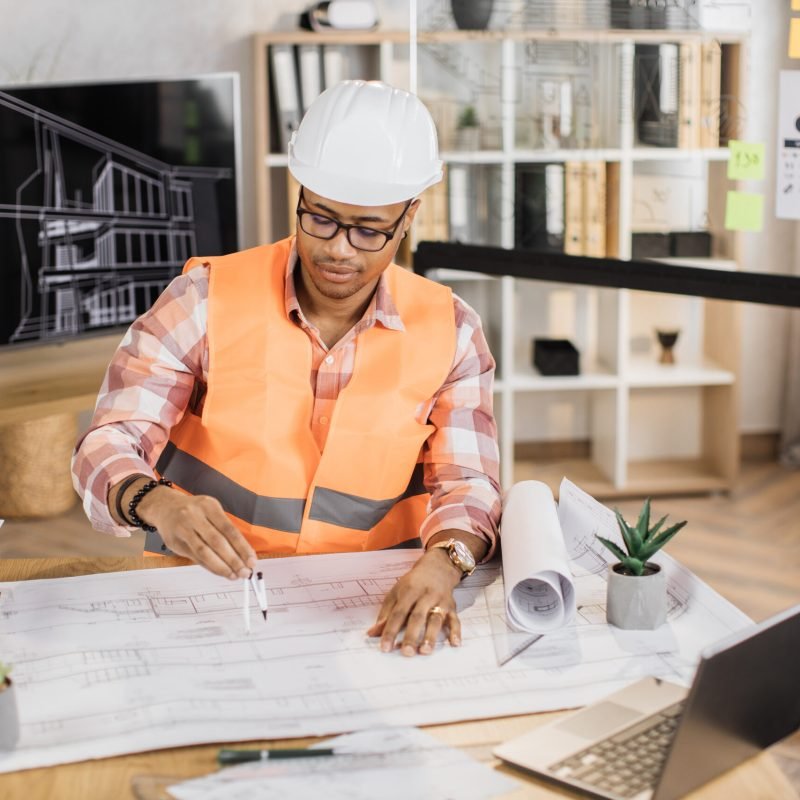 Young african builder, engineer in reflective vest uniform and hard hat on desk in office on background of large TV screen with , browsing in laptop and looking through papers or sketches.